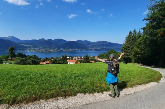 Ein Wanderer auf einem Fußweg, im Hintergrund sieht man eine Landschaft mit Dörfern, Seen und Gebirge.